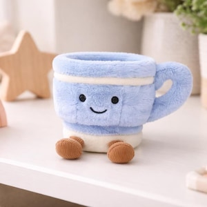 Puede incluir: Una taza de peluche azul claro con una cara sonriente, detalles blancos y pies marrones. La taza tiene un asa y está sobre una superficie blanca. Otros elementos decorativos están en el fondo.
