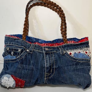 May include: A denim tote bag with a braided brown leather handle. The bag is made from repurposed jeans, featuring pockets and decorative patches. The interior lining is a red and blue paisley pattern.