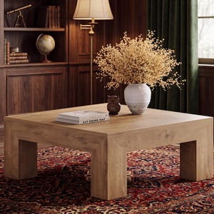 May include: A square, light brown wooden coffee table with thick, block-style legs. On the table sits a white ceramic vase filled with dried yellow flowers, a small dark brown owl figurine, and two stacked books titled ABODE and EAT DRINK NAP.