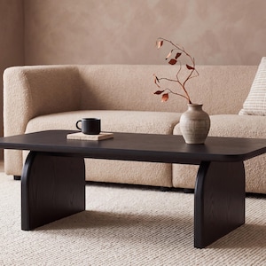 May include: A low, rectangular coffee table made of dark wood with a smooth surface and two wide, arched support legs. On the table sits a small black mug on a square coaster and a light-colored ceramic vase containing a single thin, dry branch.