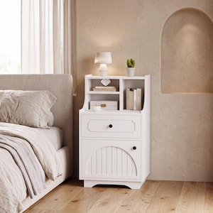 May include: A white wooden nightstand featuring an open top shelf with a small lamp, a potted cactus, and books. Below the shelf is a single drawer with a round black knob, followed by a cabinet door with a vertical slat arch design and black knob.
