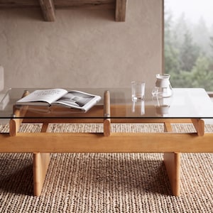 May include: A rectangular coffee table with a clear glass top and a light-toned wooden base featuring horizontal beams and vertical supports. An open book, a clear glass tumbler, and a small glass carafe are placed on the glass surface.