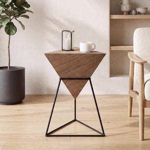May include: A side table featuring a wooden top shaped like an inverted pyramid, supported by a thin black metal frame. The table has a triangular base and a flat, wide top surface. A white candle in a black wire holder and a white mug sit on the table.