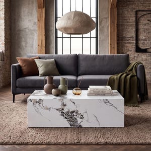 May include: A rectangular coffee table with a white surface featuring grey and black marble veining. The table is positioned in front of a dark grey sofa with brown and green cushions, a green throw blanket, and three ceramic vases.