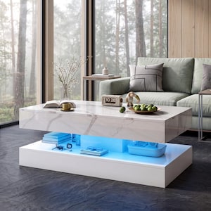 May include: A rectangular coffee table with a white marble-patterned top and a solid white base. The center section features integrated blue LED lighting that illuminates a storage shelf containing books, keys, and a small blue container.