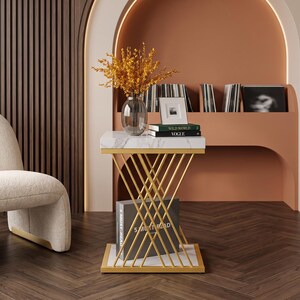 May include: A square side table with a white marble top and base, featuring a gold-colored metal frame with intersecting diagonal bars. The table holds a glass vase with yellow flowers, a small picture frame, and two books titled Wild World and Vogue.