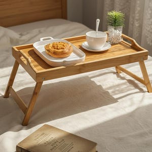 May include: A rectangular bamboo breakfast tray with foldable legs, featuring a white ceramic rectangular plate holding a golden-brown pastry, a white ceramic cup and saucer with a small spoon, and a small potted green plant in a glass vase.