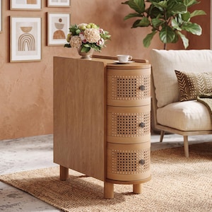 May include: A narrow, light-toned wooden side table featuring three rounded, stacked drawers with woven cane fronts and small dark circular knobs. The piece stands on four short, cylindrical legs and has a flat top surface holding a small white cup.