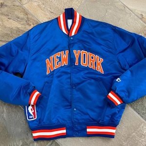 Puede incluir: Chaqueta de satén azul con las palabras "NEW YORK" en letras naranjas en el pecho. La chaqueta tiene ribetes a rayas blancas, rojas y naranjas en el cuello, los puños y la cintura. Un parche con el logo de la NBA está en la manga izquierda.
