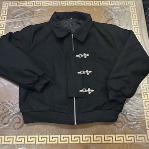 May include: A black jacket featuring a fold-down collar, long sleeves with ribbed cuffs, and a full-length front zipper. A vertical fabric panel covers the zipper, secured by three silver-toned metal toggle clasps. The interior lining is black.