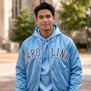 Puede incluir: Chaqueta de sat&eacute;n azul claro con la palabra "CAROLINA" en letras blancas. La chaqueta tiene una sudadera con capucha azul claro a juego debajo. La chaqueta tiene una parte delantera abotonada y ribetes blancos en la capucha y las mangas.