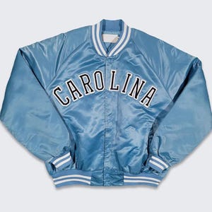 Puede incluir: Chaqueta de sat&eacute;n azul claro con la palabra "CAROLINA" en letras blancas en el pecho. La chaqueta tiene cuello, pu&ntilde;os y dobladillo acanalados con rayas blancas. Estilo universitario cl&aacute;sico.