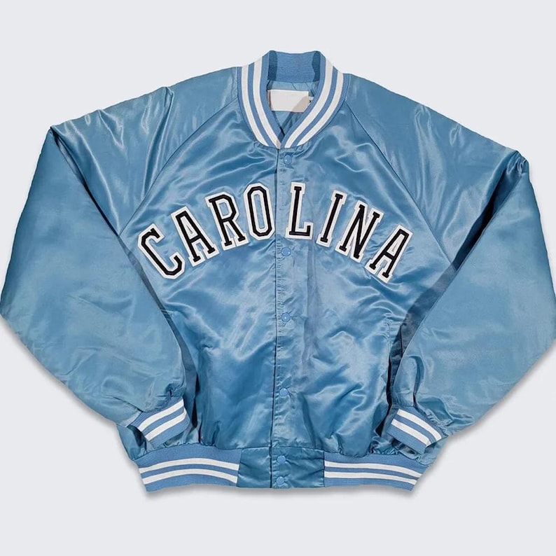 Puede incluir: Chaqueta de sat&eacute;n azul claro con la palabra "CAROLINA" en letras blancas en el pecho. La chaqueta tiene cuello, pu&ntilde;os y dobladillo acanalados con rayas blancas. Estilo universitario cl&aacute;sico.