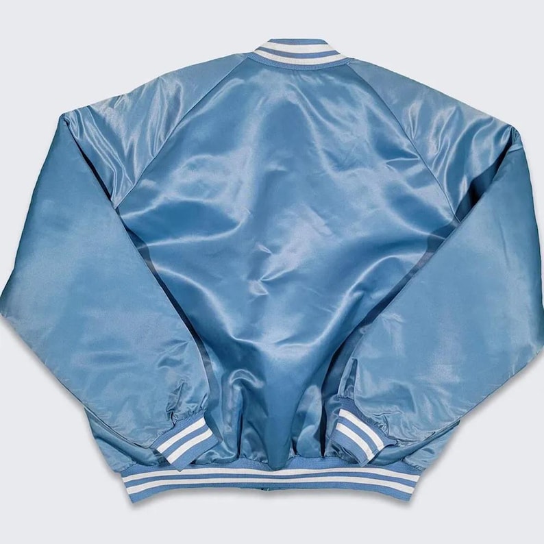Puede incluir: Chaqueta bomber de sat&eacute;n azul claro. La chaqueta presenta pu&ntilde;os, cuello y dobladillo a rayas blancas y azul claro. La chaqueta es de color liso con un acabado brillante.