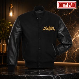 May include: A black varsity jacket featuring black leather sleeves and a black wool-blend body. The front displays the word Justice embroidered in gold thread with a cross symbol. The jacket has a snap-button closure and ribbed cuffs.