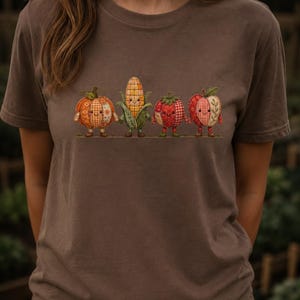 K&ouml;nnte beinhalten: Ein braunes Kurzarm-T-Shirt mit einem Grafikdruck von vier anthropomorphen Obst- und Gem&uuml;sesorten, darunter ein K&uuml;rbis, ein Maiskolben, eine Tomate und ein Apfel, die alle mit einem Patchwork-Muster und l&auml;chelnden Gesichtern gestaltet sind.