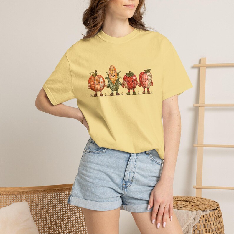 K&ouml;nnte beinhalten: Ein hellgelbes T-Shirt mit kurzen &Auml;rmeln und einem Grafikdruck von vier anthropomorphen Obst- und Gem&uuml;sesorten, darunter ein K&uuml;rbis, Mais, eine Erdbeere und ein Apfel, jeweils mit Karomuster, kleinen Gesichtern und braunen Stiefeln.