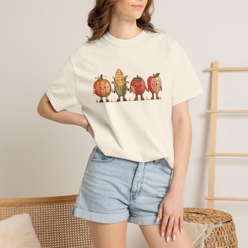 K&ouml;nnte beinhalten: Ein cremefarbenes Kurzarm-T-Shirt mit einem Grafikdruck von vier anthropomorphen Obst- und Gem&uuml;sefiguren: einem K&uuml;rbis, einem Maiskolben, einer Erdbeere und einem Apfel, die alle mit einem rot-orangen Karomuster und kleinen Gesichtern gestaltet sind.