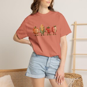 K&ouml;nnte beinhalten: Ein terrakottafarbenes Kurzarm-T-Shirt mit einem Grafikdruck von vier anthropomorphen Erntegem&uuml;sen. Das Design zeigt einen K&uuml;rbis, einen Maiskolben, eine Erdbeere und einen Apfel, alle mit einem Karomuster und kleinen Stiefeln dargestellt.