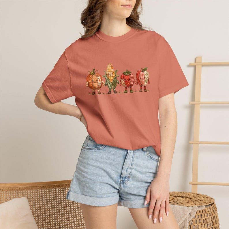 K&ouml;nnte beinhalten: Ein terrakottafarbenes Kurzarm-T-Shirt mit einem Grafikdruck von vier anthropomorphen Erntegem&uuml;sen. Das Design zeigt einen K&uuml;rbis, einen Maiskolben, eine Erdbeere und einen Apfel, alle mit einem Karomuster und kleinen Stiefeln dargestellt.