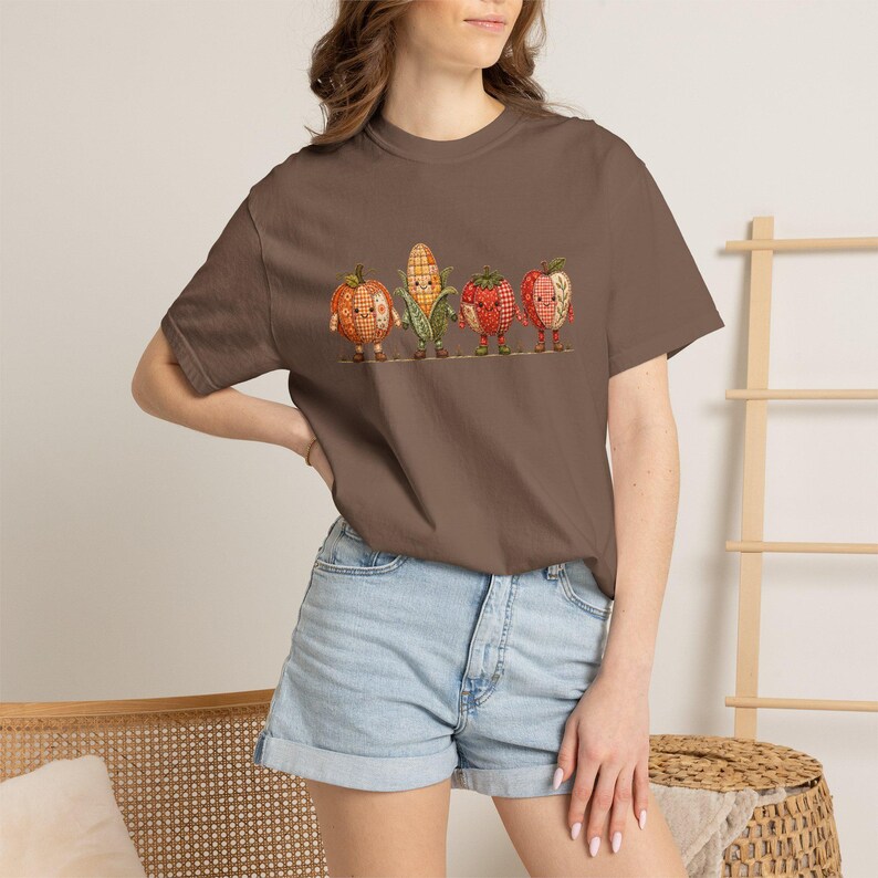 K&ouml;nnte beinhalten: Ein braunes T-Shirt mit kurzen &Auml;rmeln und einem Grafikdruck von vier anthropomorphen Obst- und Gem&uuml;sefiguren. Das Design zeigt einen K&uuml;rbis, einen Maiskolben, eine Erdbeere und einen Apfel, alle mit einem rot-orangen Karomuster versehen.