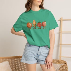 K&ouml;nnte beinhalten: Ein hellgr&uuml;nes Kurzarm-T-Shirt mit einem Grafikdruck von vier anthropomorphen Erntegem&uuml;sen und Fr&uuml;chten. Das Design zeigt einen K&uuml;rbis, einen Maiskolben, eine Erdbeere und einen Apfel, die alle mit Karomustern und kleinen Gesichtern versehen sind.