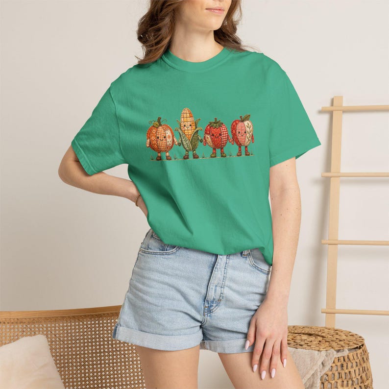 K&ouml;nnte beinhalten: Ein hellgr&uuml;nes Kurzarm-T-Shirt mit einem Grafikdruck von vier anthropomorphen Erntegem&uuml;sen und Fr&uuml;chten. Das Design zeigt einen K&uuml;rbis, einen Maiskolben, eine Erdbeere und einen Apfel, die alle mit Karomustern und kleinen Gesichtern versehen sind.