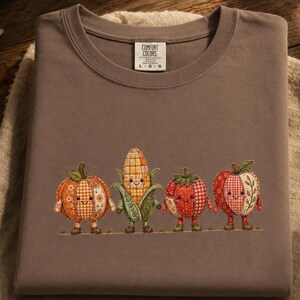 Op de afbeelding: Een taupekleurig t-shirt met ronde hals en een geborduurde afbeelding van vier antropomorfe groenten en fruit: een pompoen, een maïskolf, een tomaat en een appel, elk met een lachend gezicht en patchwork-patronen. Het label vermeldt Comfort Colors L G G.