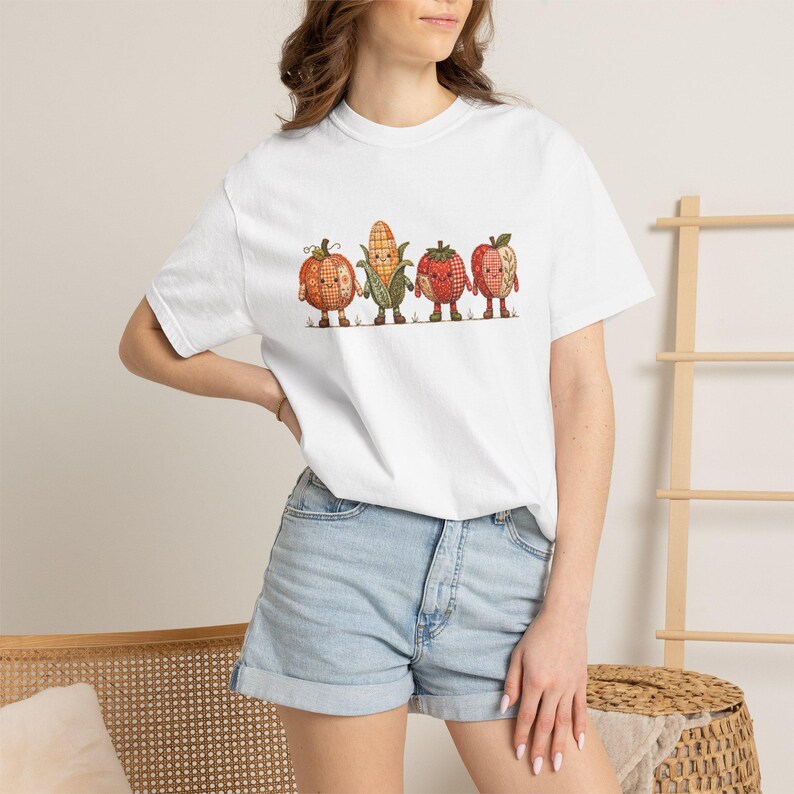 K&ouml;nnte beinhalten: Ein wei&szlig;es Kurzarm-T-Shirt mit einem Grafikdruck von vier anthropomorphen Obst- und Gem&uuml;sefiguren: einem K&uuml;rbis, einem Maiskolben, einer Erdbeere und einem Apfel, die alle mit einem Karomuster und kleinen Gesichtern gestaltet sind.