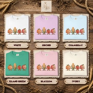K&ouml;nnte beinhalten: Sechs Rundhals-Sweatshirts in Wei&szlig;, Orchidee, Chambray, Inselgr&uuml;n, Bl&uuml;tenrosa und Elfenbein. Jedes zeigt eine Grafik von vier Cartoon-Gem&uuml;sesorten&mdash;K&uuml;rbis, Mais, Tomate und Apfel&mdash;die H&auml;ndchen halten, mit Beschriftungen unter jeder Farbe.