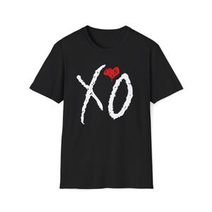May include: A black short-sleeve t-shirt featuring a white hand-drawn style XO graphic on the chest. Above the letters is a small, solid red heart shape with a textured, distressed appearance. The shirt has a standard crew neckline.