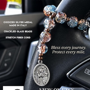 May include: A St. Christopher medal made of oxidized silver, featuring a relief of the saint carrying a child. The medal is attached to a strand of blue and clear crackled glass beads on a stretch fiber cord, with text overlaying the image.