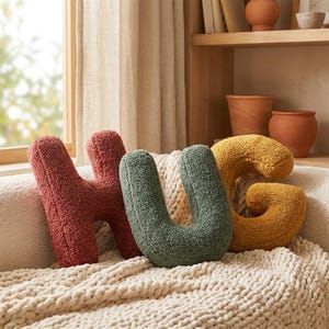 Peut inclure: Coussins décoratifs texturés épelant le mot "HUG". La lettre "H" est rouge orangé, le "U" est vert et le "G" est jaune. Les coussins sont sur une couverture crème, avec une fenêtre et des étagères en arrière-plan.