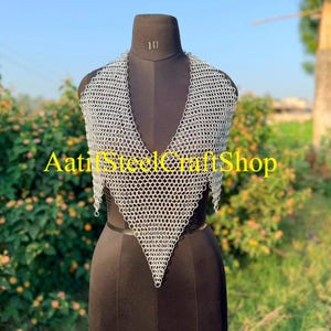 May include: A silver-colored chainmail halter top constructed from interlocking metal rings in a V-neck design. The garment is displayed on a black mannequin torso. The text AatifSteelCraftShop is overlaid in yellow across the center of the piece.