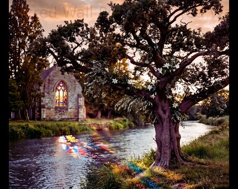 May include: A landscape wall print featuring a stone church with a glowing stained glass window beside a river. A large tree with white flowers stands on the grassy bank, reflecting colorful light onto the water and grass at sunset.