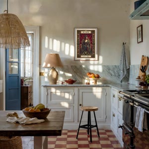 Op de afbeelding: Een keukeninterieur met een ingelijste kunstprint van het sterrenbeeld Schorpioen. De keuken heeft een houten tafel met een fruitschaal, een houten kruk en een vintage fornuis. De vloer heeft een rood-wit geblokt patroon. Een lamp staat op het aanrecht.