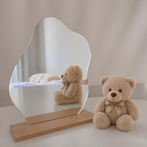 Peut inclure: Un miroir décoratif de forme organique, posé sur une base en bois, reflète un ours en peluche. Un deuxième ours en peluche est assis à droite du miroir. Les deux ours sont brun clair avec des nœuds beiges.