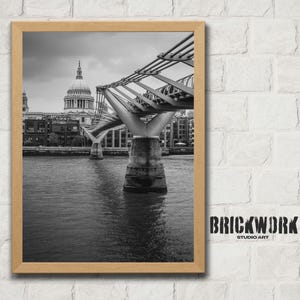 May include: Black and white framed print of the Millennium Bridge in London, with St. Paul's Cathedral in the background. The bridge spans the River Thames. The print is in a wooden frame. The text "BRICKWORK STUDIO ART" is in the lower right corner.