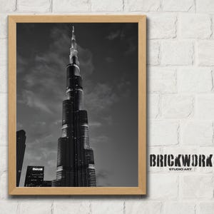Puede incluir: Impresión enmarcada en blanco y negro del rascacielos Burj Khalifa en Dubái. La imagen está ambientada contra un cielo nublado. El marco es de color madera clara. El texto "BRICKWORK STUDIO ART" está impreso en negro a la derecha.