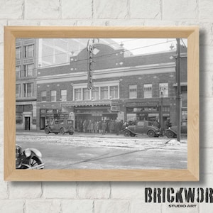 Könnte beinhalten: Gerahmter Schwarz-Weiß-Druck einer Vintage-Straßenszene mit klassischen Autos und einem Theater. Das Bild fängt einen Moment in der Zeit ein und zeigt Architektur und Fahrzeuge aus einer vergangenen Ära. Der Rahmen ist hellbraun.