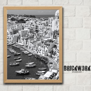 May include: Black and white framed print of a coastal town with boats in the harbor. The image features a detailed view of buildings, a harbor filled with boats, and a clear sky. The print is in a wooden frame. The text "BRICKWORK STUDIO ART" is in the lower right corner.