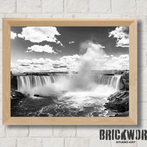 May include: A monochrome photograph of Niagara Falls, presented in a light wood frame. The image shows the waterfall, mist, and a cloudy sky. The bottom right corner of the frame has the text "BRICKWORK STUDIO ART".