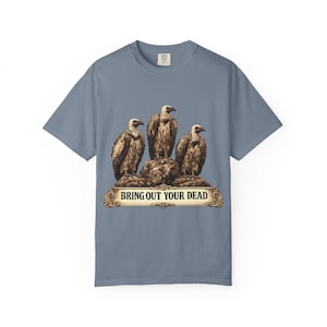 May include: A blue t-shirt featuring a graphic of three vultures perched on a rock. Below the vultures is a banner with the text "BRING OUT YOUR DEAD" in a decorative font. The shirt is a solid blue color.