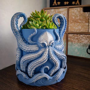 May include: A blue octopus-shaped planter with silver accents, holding a green succulent plant. The planter features detailed tentacles and a wave-like base, creating a unique decorative piece.