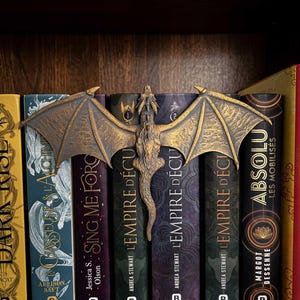 May include: A bronze-colored dragon bookend with outstretched wings rests atop a row of books. The bookend is detailed with scales and a textured surface. The books have titles like "L'Empire D'Écu" and "Sing Me Forc". The books are on a dark wooden shelf.