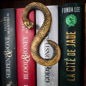 Könnte beinhalten: Ein goldener, schlangenförmiger Lesezeichen liegt zwischen Büchern. Der Lesezeichen hat ein detailliertes Schuppenmuster. Die Buchrücken sind schwarz, rot, weiß und grün, mit Titeln wie "Götter & Monster".