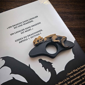 Op de afbeelding: Een zwarte, draakvormige boekenhouder met een goudkleurig drakenbeeldje erop. De boekenhouder ligt op een boek met Franse tekst. Het boek heeft een zwart draak silhouet op de omslag.
