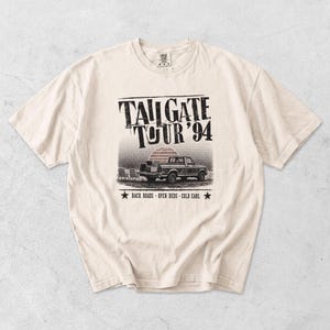May include: Cream-colored t-shirt with a distressed graphic. The graphic features a pickup truck, a sunset, and the text "TAILGATE TOUR '94". Below the image are the words "BACK ROADS - OPEN BEDS - COLD CANS".