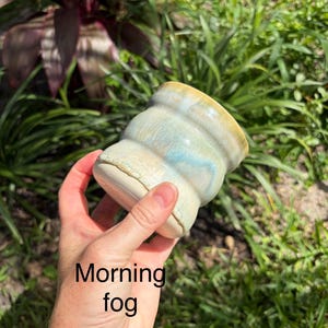 May include: A ceramic mug with a ribbed, cylindrical body featuring a glaze of blended light blue, cream, and tan tones. The surface has a smooth, glossy finish with subtle color variations. The text Morning fog is printed on the lower section.