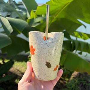 May include: A speckled cream-colored ceramic tumbler featuring small orange and brown leaf motifs. The vessel has a notched rim designed to hold a straight, silver-colored metal straw that extends vertically from the top opening.